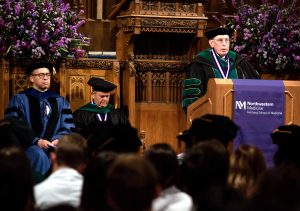 Man in formal academic regalia speaks at a potium with a purple banner draped over it. Individuals in similar regalia sit behind him. in the foreground are the backs of people sitting in rows.