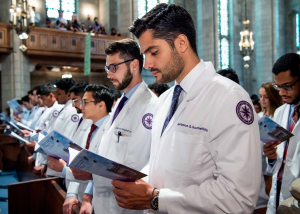 Rows of people in white coats with purple patches read from booklets. The setting is a church.