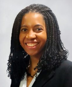 Woman smiling. She has long braids and wears a black suit jacket and white blouse.